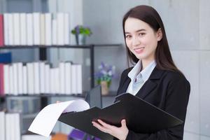 Asian business woman in black suit smiles happily while she works and holds clipbroad in office. photo