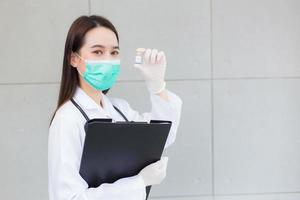 Asian woman doctor who wears medical face mask holds document file in her hand and another holds vaccine COVID-19 vaccine bottle to protect health from pathogen epidemic. photo