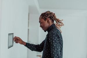 Smiling African American man using modern smart home system, controller on wall, positive young man switching temperature on thermostat or activating security alarm in apartment photo