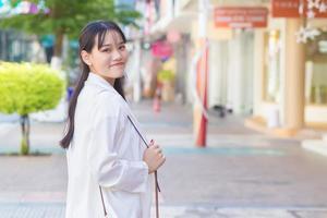 una joven trabajadora asiática de negocios segura de sí misma que usa una camisa blanca y una bolsa de hombro sonríe alegremente mientras camina para trabajar en la oficina en la ciudad con un edificio de tiendas en el fondo. foto