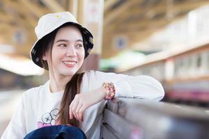 Asian beautiful girl in a long-sleeved white shirt and a hat sits happy smilie in the train station waiting for the train to arrive.