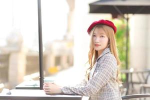 Asian beautiful girl who wears suit and red cap with bronze hair sits on chair in coffee shop while there was a coffee mug on the table on a sunny morning. photo