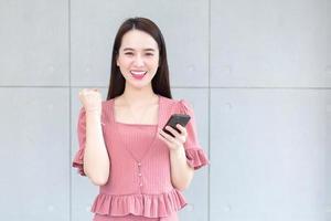 Asian beautiful woman in a pink dress smiles happily successful while holds smartphone. photo