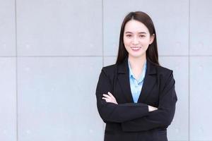 mujer trabajadora asiática que tiene el pelo largo viste traje negro formal con camisa azul mientras cruza el brazo y sonríe alegremente. foto