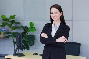 Asian working woman who has long hair wears black formal suit with blue shirt while she arm crossing and smiling happily. photo