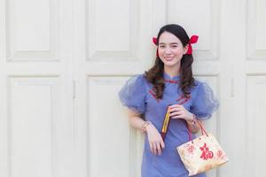 una chica asiática con un vestido chino gris azulado sostiene la bolsa de papel que proyecta la palabra que significa feliz en chino y se para en una puerta de madera blanca como fondo. foto
