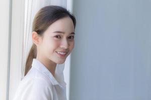 hermosa mujer asiática tiene pelo largo negro en camisa blanca. ella está sonriendo y parada cerca de la ventana con una cortina blanca. foto