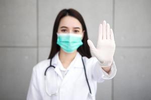 Asian woman doctor who wears medical coat and face mask shows hand as stop sign in health protection concept at hospital. Stop epidemic of COVID-19. photo