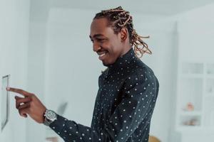 Smiling African American man using modern smart home system, controller on wall, positive young man switching temperature on thermostat or activating security alarm in apartment photo