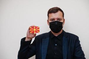 el hombre europeo usa negro formal y protege la máscara facial, sostiene la tarjeta de la bandera de macedonia del norte aislada en el fondo blanco. concepto de país covid coronavirus de europa. foto