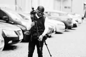 Young professional african american videographer holding professional camera with tripod pro equipment. Afro cameraman wearing black duraq making a videos. photo