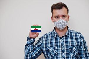 Man in checkered shirt show Federal Dependencies of Venezuela flag card in hand, wear protect mask isolated on white background. American countries Coronavirus concept. photo