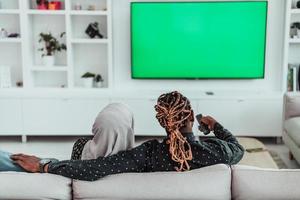 African Couple Sitting On Sofa Watching TV Together Chroma Green Screen Woman Wearing Islamic Hijab Clothes photo