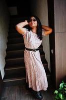 Fashion portrait of african american woman in long dress, boots and sunglasses indoor. photo