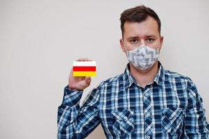 Man in checkered shirt show South Ossetia flag card in hand, wear protect mask isolated on white background. Europe countries Coronavirus concept. photo