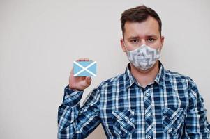 Man in checkered shirt show San Andrs and Providencia flag card in hand, wear protect mask isolated on white background. American countries Coronavirus concept. photo