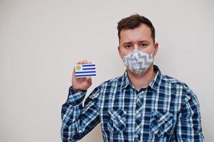 Man in checkered shirt show Uruguay flag card in hand, wear protect mask isolated on white background. American countries Coronavirus concept. photo