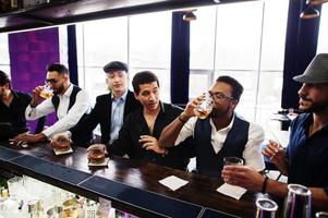 Group of handsome retro well-dressed man gangsters spend time at club. Drinking whiskey at bar counter. Multiethnic male bachelor mafia party in restaurant. photo