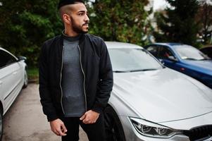 retrato de un elegante hombre de barba árabe con cuello alto gris y chaqueta negra. chico modelo árabe parado cerca de su coche. foto