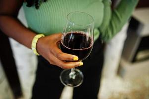 Cierra las manos de una atractiva joven afroamericana vestida con suéter verde y jeans negros posan en el restaurante con una copa de vino tinto. foto
