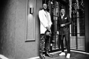dos hombres negros de moda posan contra la casa con guirnaldas. retrato de moda de modelos masculinos afroamericanos. llevar traje, abrigo y sombrero. foto