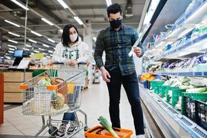 Asian couple wear in protective face mask shopping together in supermarket during pandemic. Taking vegetables from fridge. photo