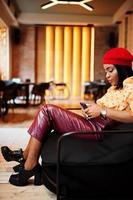 Elegant afro american woman in red french beret, big gold neck chain polka dot blouse and leather pants sit on pouf with mobile phone. photo