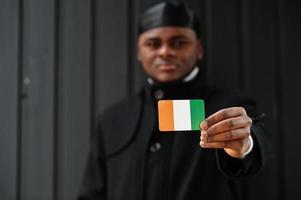 African man wear black durag hold Ivory Coast flag at hand isolated dark background. photo