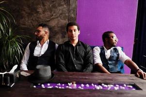 Group of handsome retro well-dressed man gangsters spend time at club. Multiethnic male bachelor mafia party in restaurant, sitting by table. photo