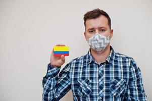 Man in checkered shirt show Colombia flag card in hand, wear protect mask isolated on white background. American countries Coronavirus concept. photo