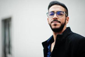 Middle eastern entrepreneur wear black coat and blue shirt, eyeglasses standing against white wall. photo