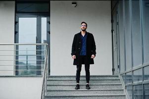 Middle eastern entrepreneur wear black coat and blue shirt, eyeglasses against office building, stand on stairs. photo