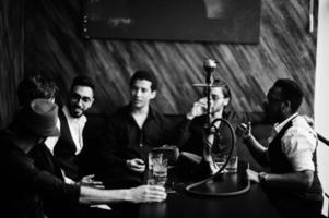 Group of handsome retro well-dressed man gangsters spend time at club, smoking hookah. Multiethnic male bachelor mafia party in restaurant. photo