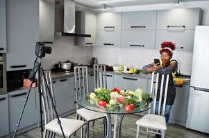 African american woman filming her blog broadcast about healthy food at home kitchen and looking at phone. photo