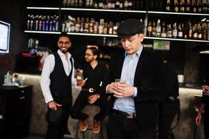 Stylish arab man against group of handsome retro well-dressed guys gangsters spend time at club, drinking on bar counter. Multiethnic male bachelor mafia party in restaurant. photo