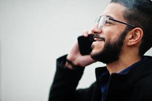 Middle eastern entrepreneur wear black coat and blue shirt, eyeglasses standing against white wall, smoking cigarette and speak by phone. photo