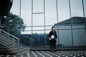 Middle eastern entrepreneur wear black coat and blue shirt, eyeglasses against office building with laptop. photo