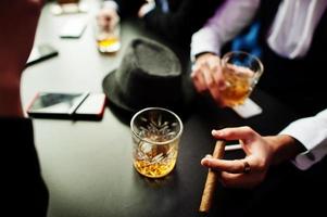 Group of handsome retro well-dressed man gangsters spend time at club, sitting and drinking whiskey. Multiethnic male bachelor mafia party in restaurant. Hand of man with cigar. photo