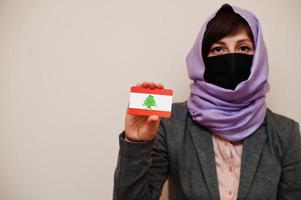 retrato de una joven musulmana que usa ropa formal, protege la máscara facial y el pañuelo de la cabeza del hiyab, sostiene la tarjeta de la bandera del líbano contra un fondo aislado. concepto de país coronavirus. foto