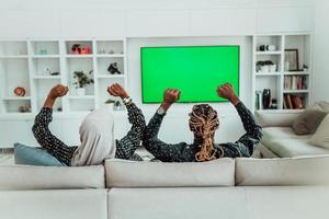 African Couple Sitting On Sofa Watching TV Together Chroma Green Screen Woman Wearing Islamic Hijab Clothes photo