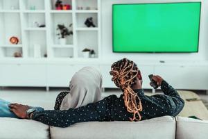 African Couple Sitting On Sofa Watching TV Together Chroma Green Screen Woman Wearing Islamic Hijab Clothes photo