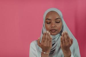 mujer musulmana africana moderna hace oración tradicional a dios, mantiene las manos en gesto de oración, usa ropa blanca tradicional, tiene expresión facial seria, aislada sobre fondo rosa plástico foto