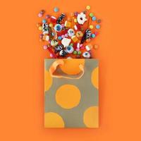 Shopping paper orange polka dot gift bag full of spilled assorted traditional Halloween candies. Orange square background with copy space. photo