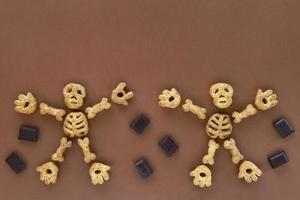 Puffed corn snacks Halloween treat in skeleton form on brown background. Dancing skeleton figures made of parts - skull, rib cage, hands, feet, bones. Pieces of chocolate around. photo