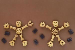 Puffed corn snacks Halloween treat in skeleton form on brown background. Dancing skeleton figures made of parts - skull, rib cage, hands, feet, bones. Pieces of chocolate around. photo