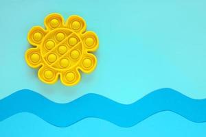 Yellow sun new trendy anti stress sensory silicone bubble fidget toy on blue seascape background, paper cut waves and sky. Pop it toy for relaxation on summer sea vacation. Place for text. photo