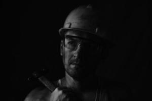 Miner, dirty in coal soot, wearing helmet, protective glasses and with hammer in his hands in black and white photo. photo