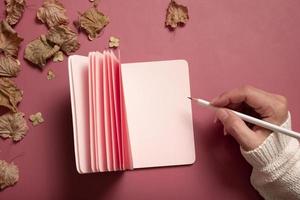 Office table desk with blank pink paper note pad and female hand wit pencil and fall leaves on colored background. Autumn flat lay composition with blank paper photo