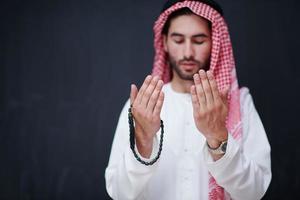 arabian man making traditional prayer to God, keeps hands in praying gesture in front of black chalkboard photo