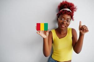 mujer africana con cabello afro, use camiseta amarilla y anteojos, sostenga la bandera de guinea aislada en fondo blanco, muestre el pulgar hacia arriba. foto